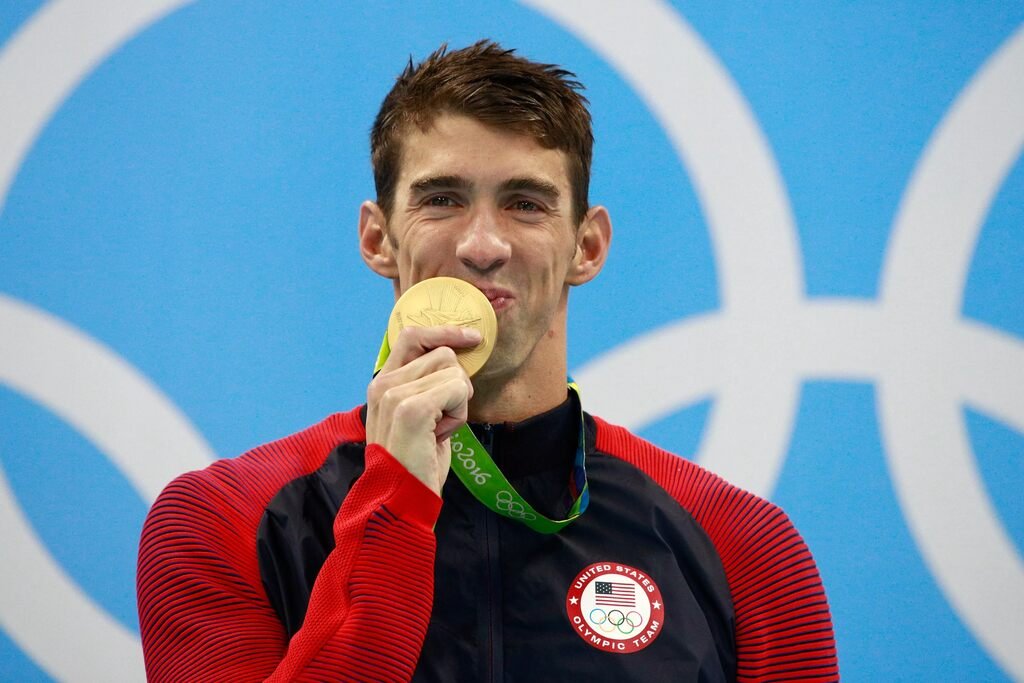 Na imagem, o nadador olímpico Michael Phelps é visto mordendo sua medalha de ouro durante a cerimônia de premiação nas Olimpíadas de 2016, no Rio de Janeiro. Ele está vestindo uma jaqueta vermelha e preta com o emblema da equipe dos Estados Unidos e exibe um sorriso de vitória e satisfação. O fundo azul claro, com os anéis olímpicos visíveis, completa a cena de celebração.