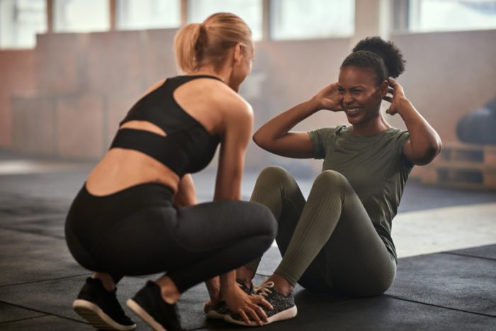 Duas mulheres fazendo atividade física, mais precisamente uma ajudando a outra no abdominal. 